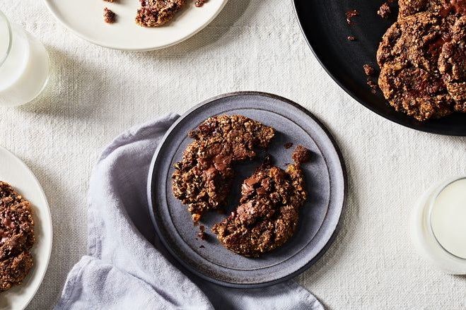 A dark brown, crumbled in half chocolate chip cookie sits on a gray plate with several other plates on a white table cloth.